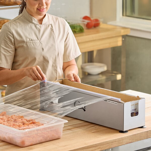 A stainless steel film and foil dispenser and cutter being used to cover a food container with plastic wrap.
