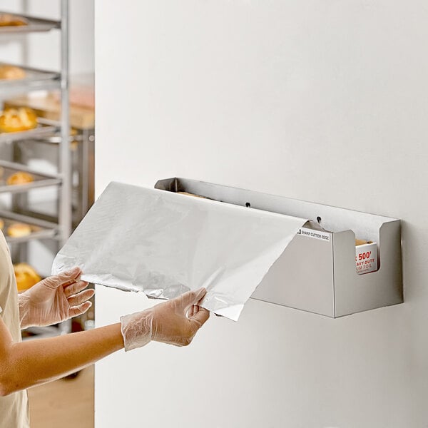 A stainless steel film and foil dispenser and cutter mounted on a wall, with a person pulling out a sheet of foil.