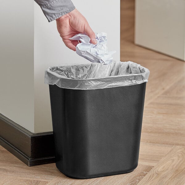 A hand throwing a piece of paper into a black Rubbermaid fire-resistant rectangular wastebasket.