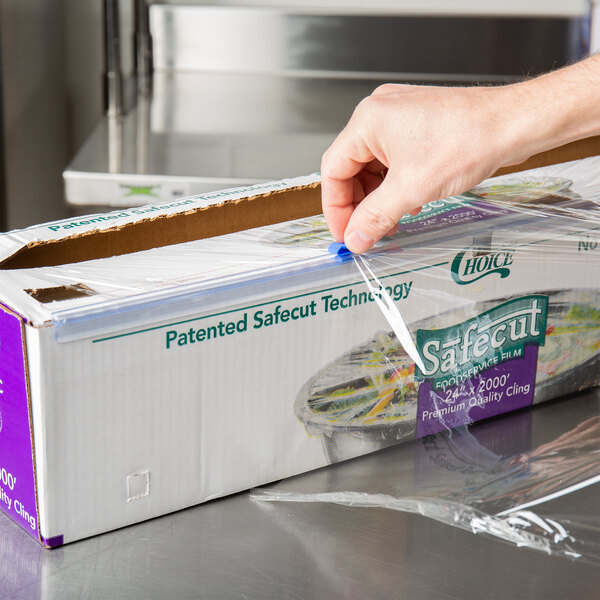 A hand using a Choice 24" safety slide cutter to cut plastic wrap over a box.