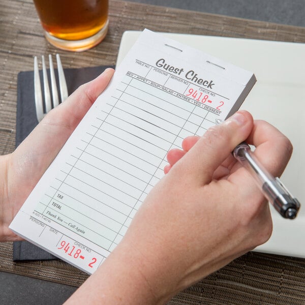 A person holding an Adams guest check book and writing with a pen.