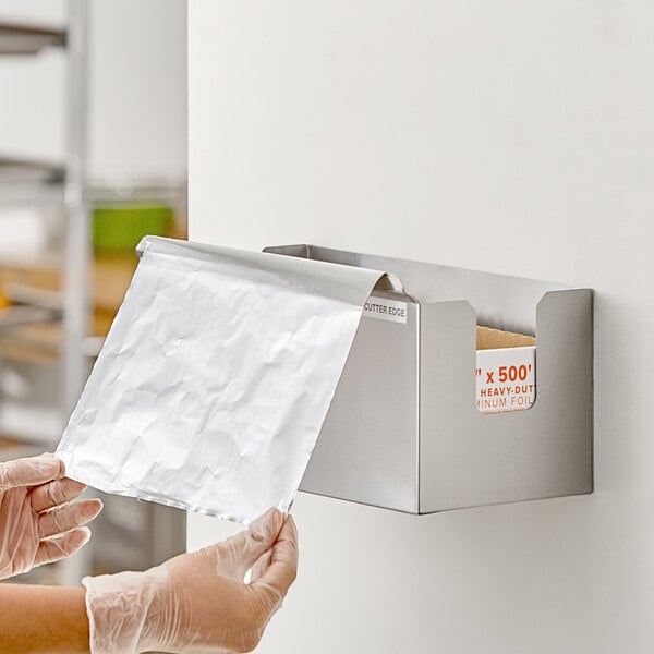 A stainless steel dispenser and cutter for film and foil mounted on a wall, with a sheet of aluminum foil being pulled out.