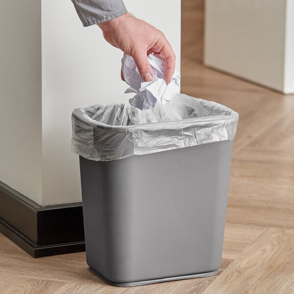 A hand holding crumpled paper over a grey Continental rectangular wastebasket with a plastic bag inside.