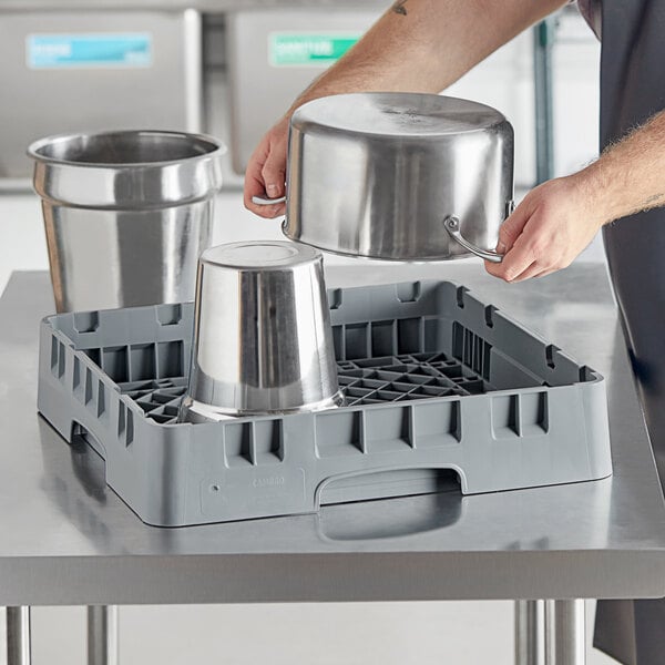 A hand holding a silver pot over a grey plastic Cambro dish rack.