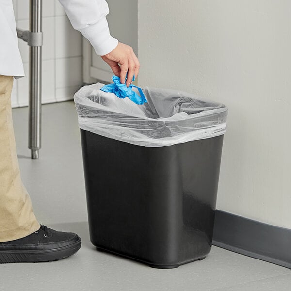 A person putting a plastic bag in a black Rubbermaid rectangular wastebasket.