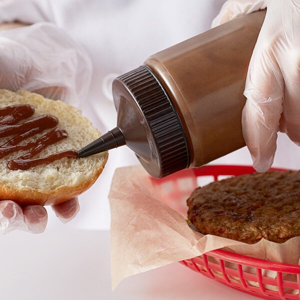 A person using a Vollrath brown wide mouth squeeze bottle to put sauce on a hamburger bun.