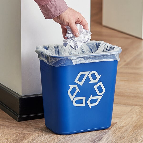 A hand putting a crumpled paper into a blue Rubbermaid recycling bin.