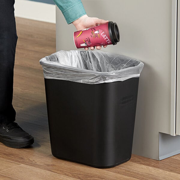 A hand putting a coffee cup into a Rubbermaid black rectangular wastebasket.
