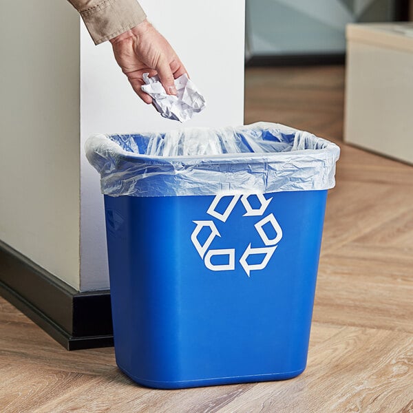 A hand throwing crumpled paper into a blue Rubbermaid recycling bin.