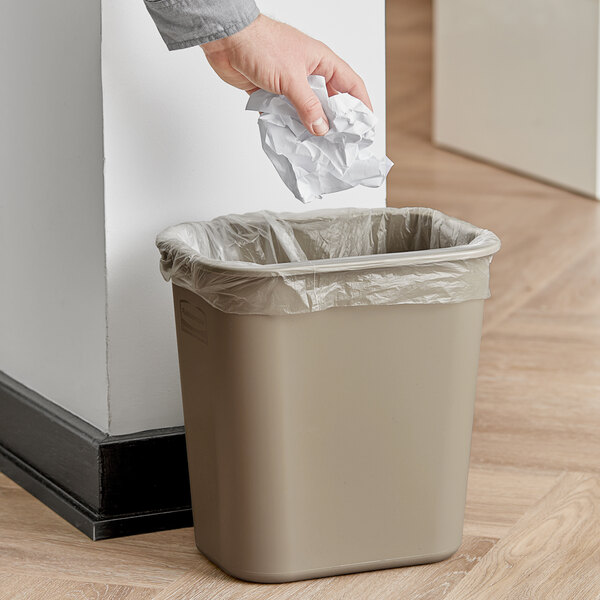 A hand holding a piece of paper and putting it into a Rubbermaid beige rectangular trash can with a plastic bag inside.