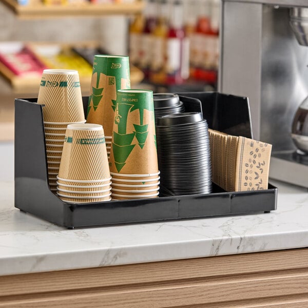 A black three-section countertop organizer holding disposable cups, lids, and napkins.