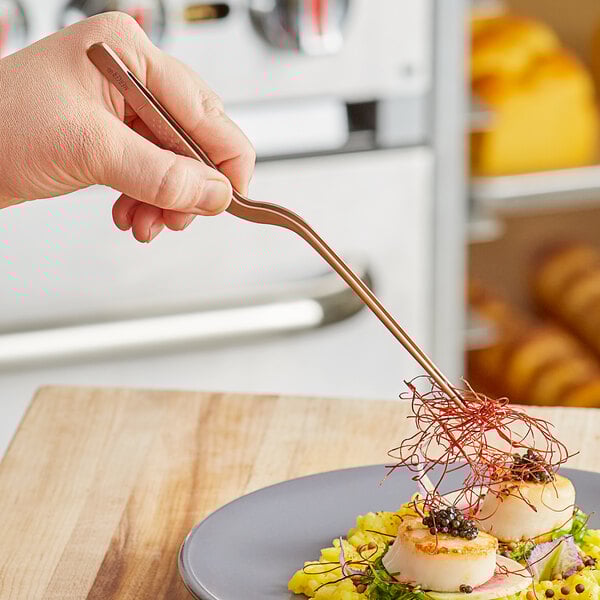A hand using Mercer Culinary rose gold offset tip plating tongs to plate food.