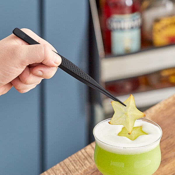 Mercer Culinary black straight tip plating tongs holding a star fruit on top of a drink.