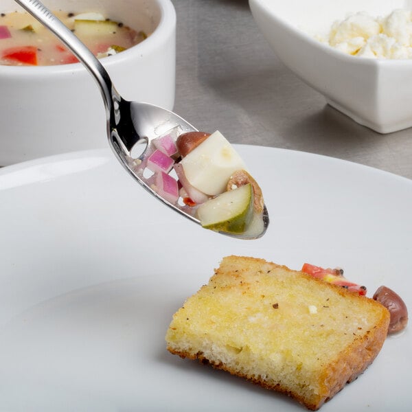 A Mercer Culinary stainless steel slotted bowl plating spoon holding a piece of bread over a bowl of soup.