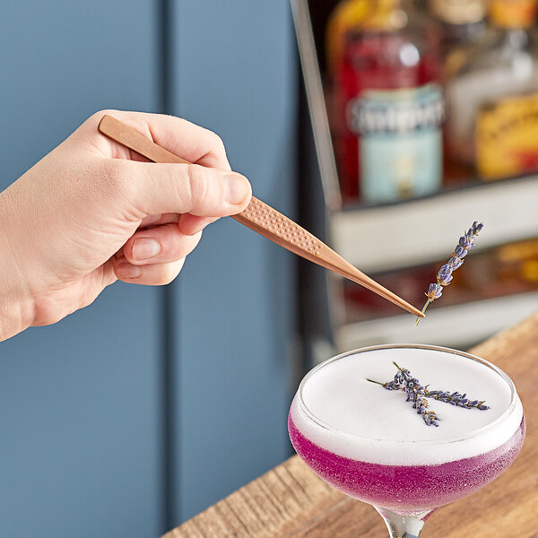 A person using Mercer Culinary rose gold plating tongs to garnish a purple cocktail.