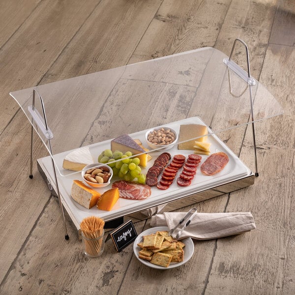 A rectangular Frilich portable sneeze guard over a table of food with crackers, cheese and grapes.