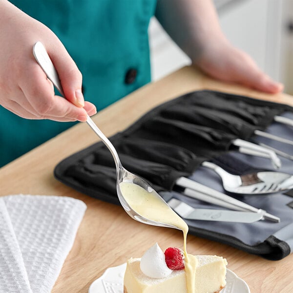 A person using a Mercer Culinary stainless steel spoon to pour white sauce on a slice of cake.