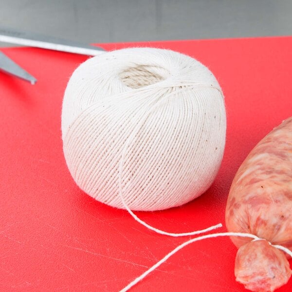 Roll of white cotton twine next to meat and scissors on red cutting board
