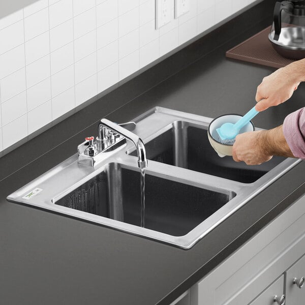 A person washing dishes in a Regency stainless steel drop-in sink with running water.