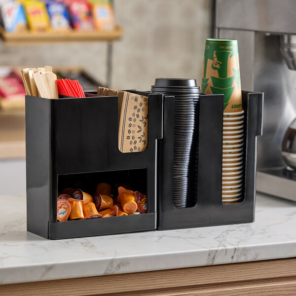 A black six-section countertop organizer holding cups, lids, napkins, stirrers, and coffee pods.