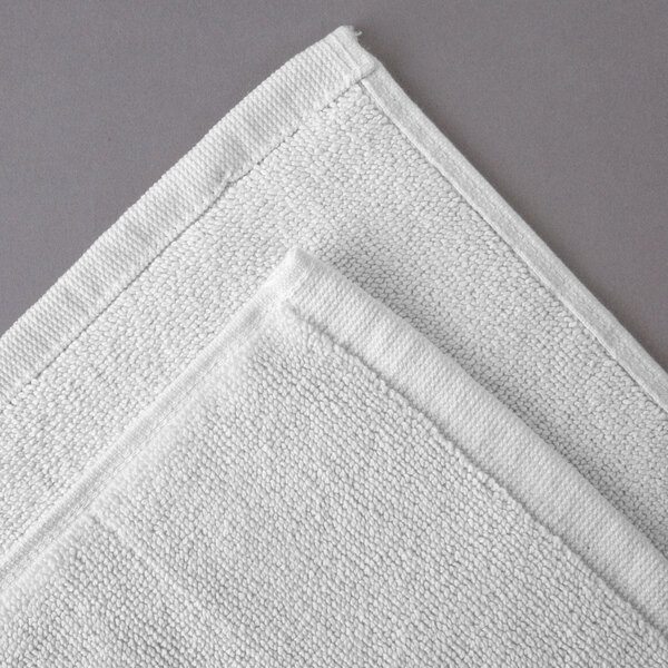 An Oxford Vicenza Bianco white bath mat with a Dobby border on a gray surface.