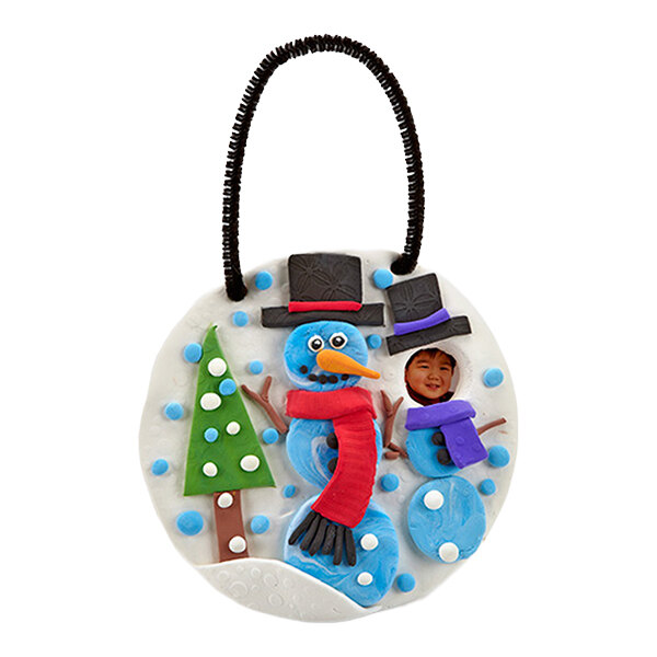 A round ornament featuring a snowman and a child’s face incorporated into a smaller snowman, with a green tree and blue and white decorations, and a black loop for hanging.