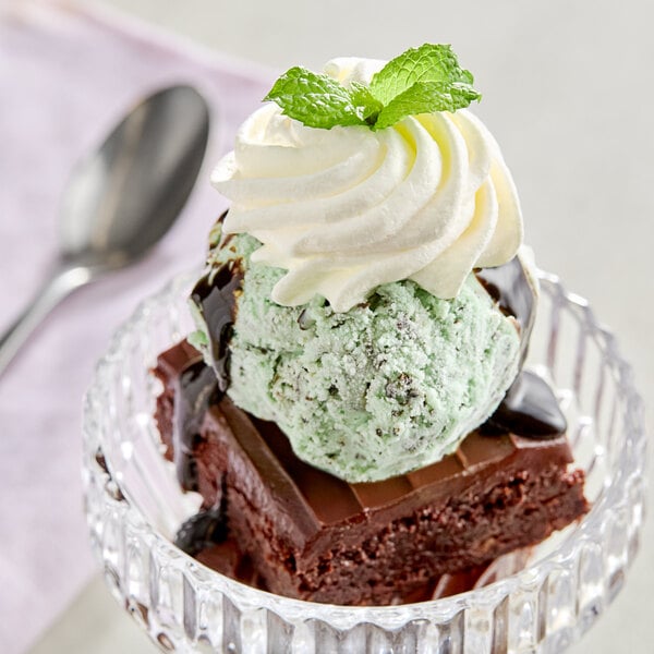 A dessert featuring a brownie topped with mint chocolate chip ice cream, whipped cream, chocolate sauce, and a mint leaf, served in a glass dish.
