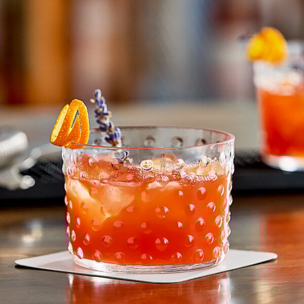 A close up of an Acopa hobnail rocks glass filled with orange liquid and a slice of orange.