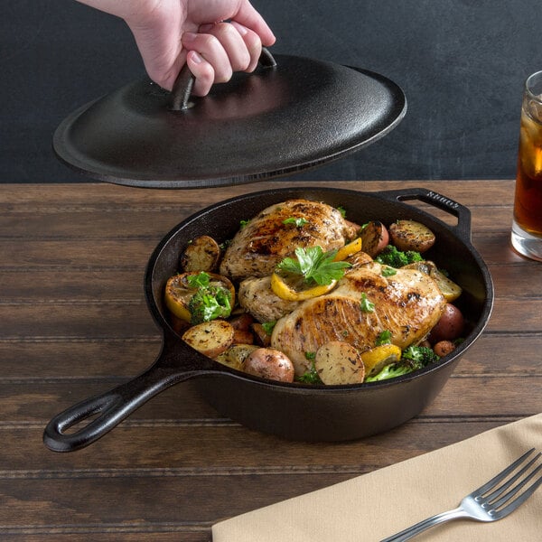 A hand holding a Lodge cast iron lid over a pan of chicken and potatoes.