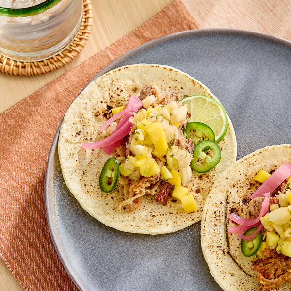 A plate with two tacos topped with shredded meat, pineapple salsa, pickled onions, and sliced jalapeÃ±os, garnished with lime wedges.
