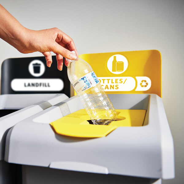 A hand putting a plastic bottle into a Rubbermaid recycling bin with a bottle lid.