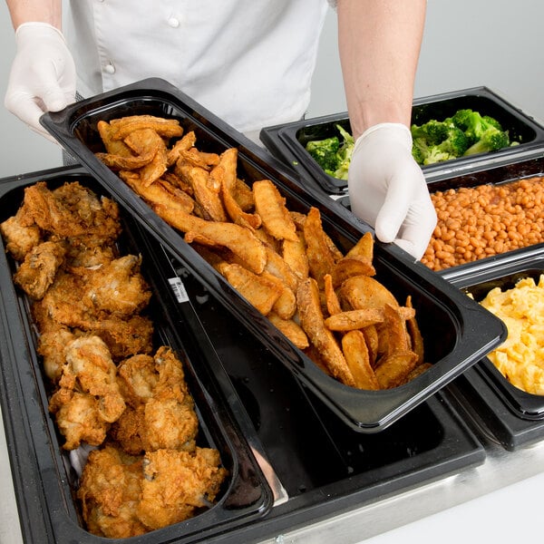 A person in white gloves holding a Cambro black plastic food pan full of food.