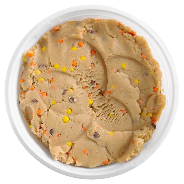 A round container filled with light brown cookie dough that has orange, yellow, and brown candy pieces mixed in.