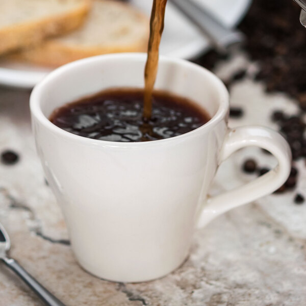 A close-up of coffee being poured into a Tuxton Europa white cappuccino mug.