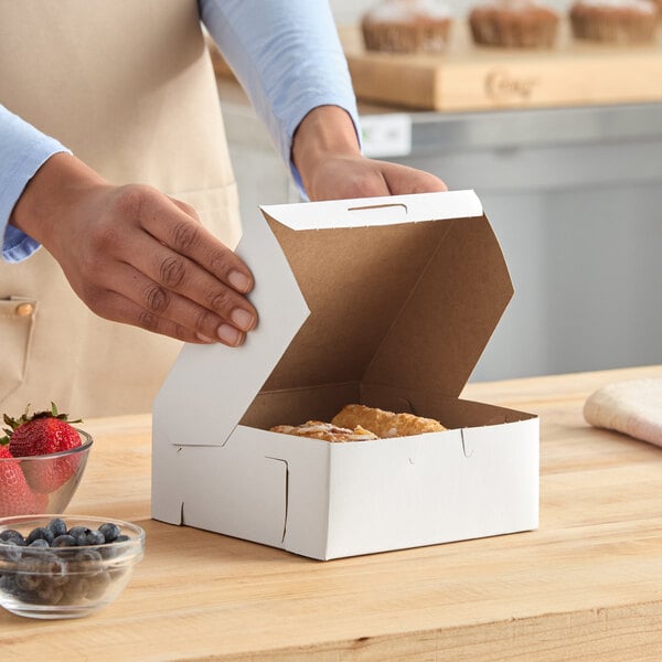 A white, customizable 6 x 6 x 2.5 inch bakery box being assembled on a wooden counter.