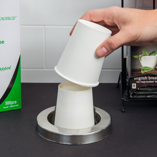 A hand using a San Jamar black plastic adjustment ring to hold a white cup over a coffee cup dispenser.