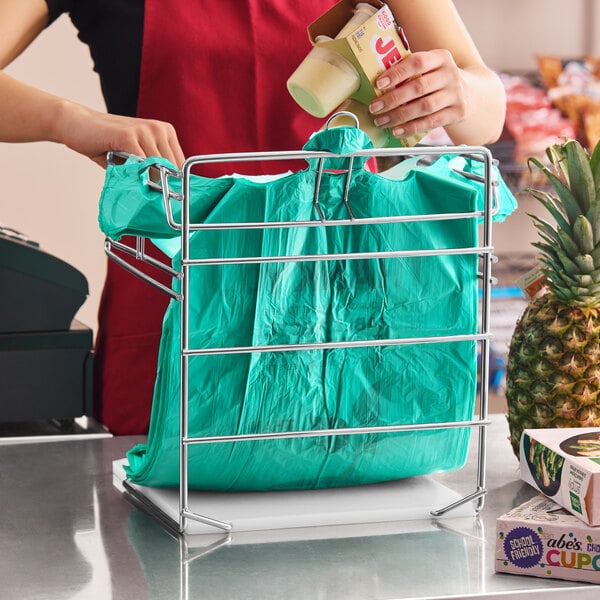 A chrome t-shirt bag rack stand holding a green plastic shopping bag at a checkout counter.
