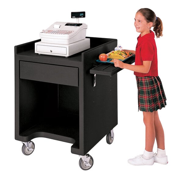 A girl in a red sweater and plaid skirt standing next to a black Cambro cash register stand with trays on both sides.