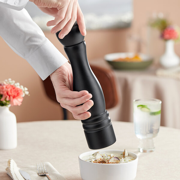 A person using a black Acopa wooden pepper mill to season a bowl of soup.