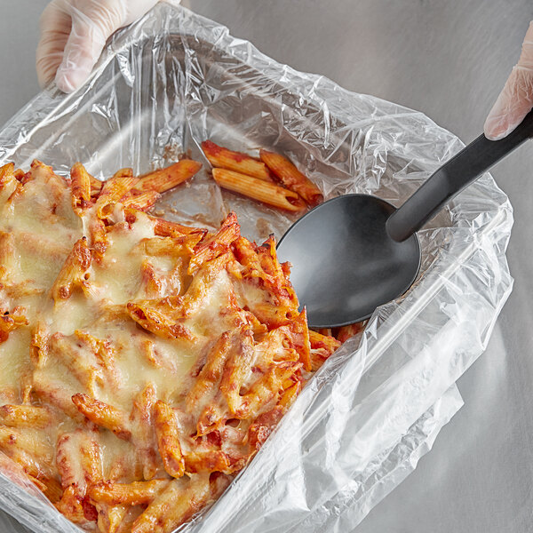 A hand using a black ladle to serve pasta from a stainless steel pan with a plastic liner.