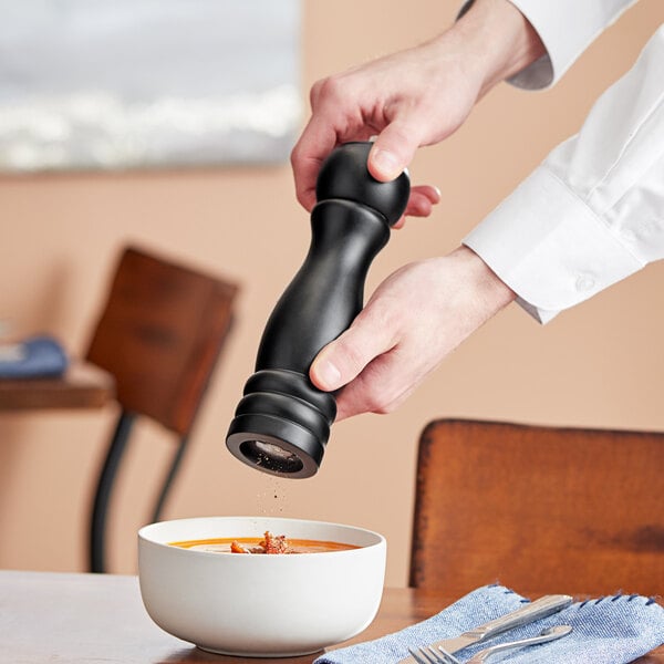 A hand using a black Acopa pepper grinder to season a bowl of soup.