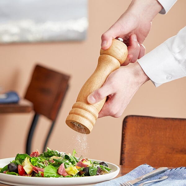 A person using an Acopa wooden pepper mill to sprinkle pepper over a salad.