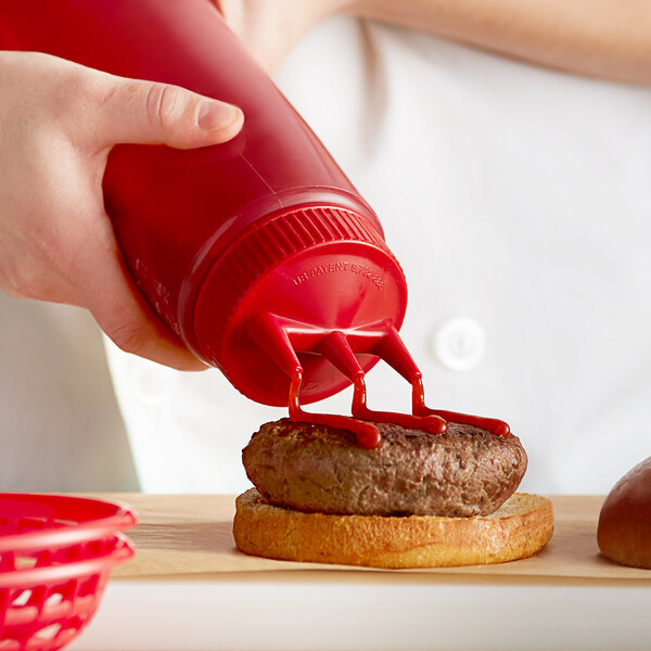 A hand using a Vollrath red Tri Tip squeeze bottle to pour ketchup onto a burger.