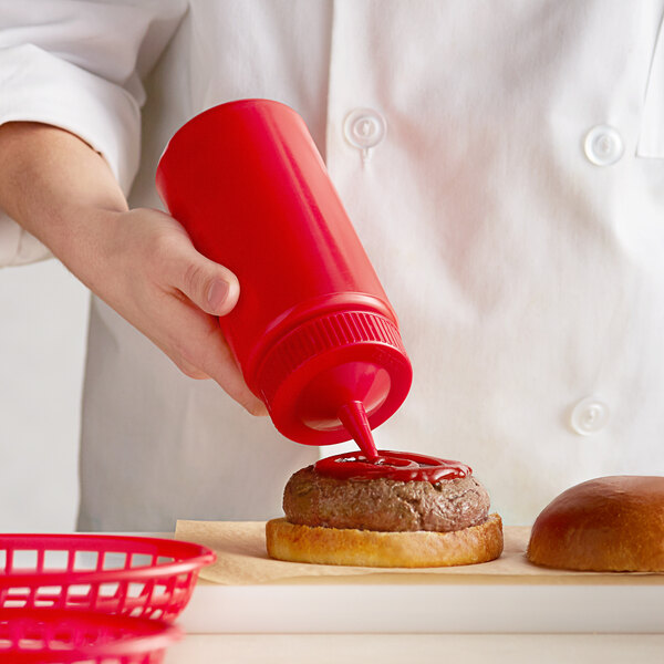 A hand using a Vollrath red wide mouth squeeze bottle to pour ketchup on a hamburger.