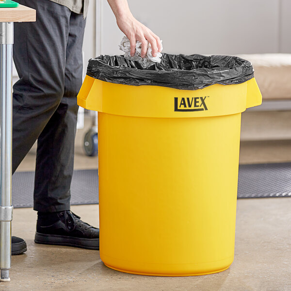 A man putting a garbage bag into a yellow Lavex trash can.