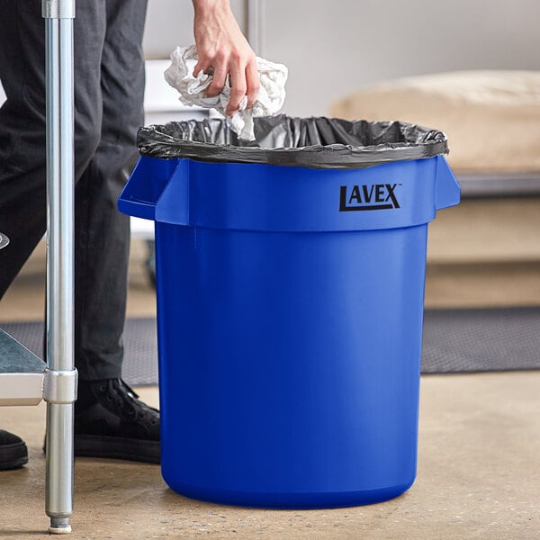 A man putting a black plastic bag in a blue Lavex commercial trash can.