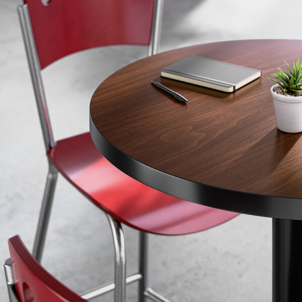 A round bar-height table with a walnut finish, accompanied by red chairs and a small potted plant on top.