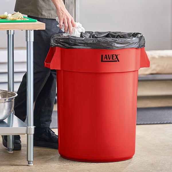 A man wearing black pants and a black shirt putting a bag in a red Lavex trash can.