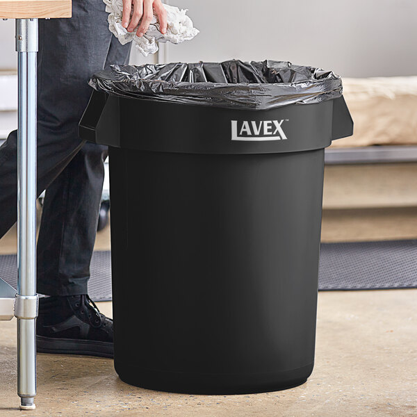 A man putting a black garbage bag in a Lavex 32 gallon black round commercial trash can.