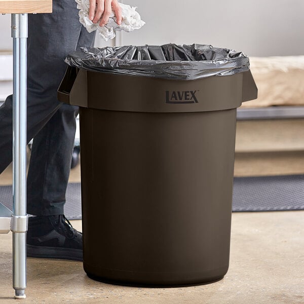 A man standing next to a brown Lavex commercial trash can with a white object inside.